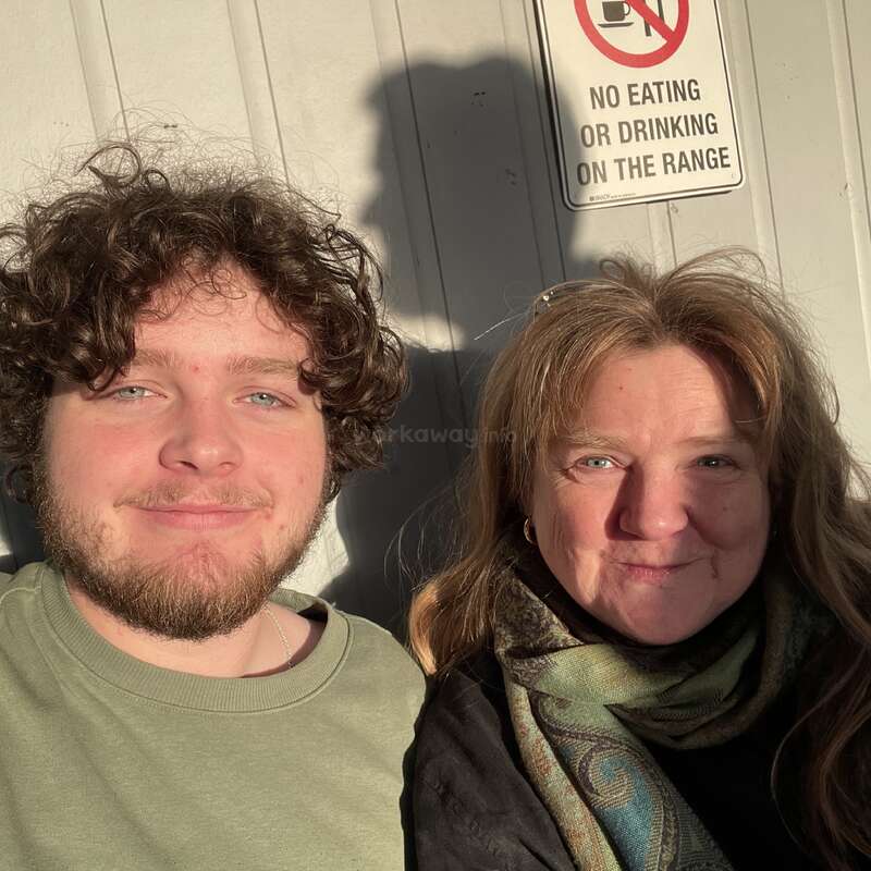 Um rapaz e uma moça sorriem sob a luz quente do sol, parados ao lado de uma parede com uma placa "Proibido comer ou beber no campo" acima de suas cabeças.