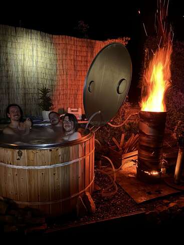 Drei Typen chillen nachts in einem Holz-Whirlpool. In der Nähe gibt's einen selbstgebauten Ofen, der eine hohe, helle Flamme macht und so eine gemütliche und rustikale Atmosphäre schafft.