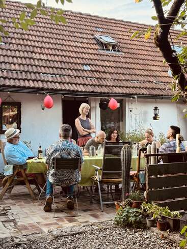 Eine Gruppe Leute sitzt draußen an einem Tisch und genießt gemeinsam ein Essen neben einem rustikalen Haus. Luftballons und Pflanzen sorgen für eine fröhliche, entspannte Atmosphäre.