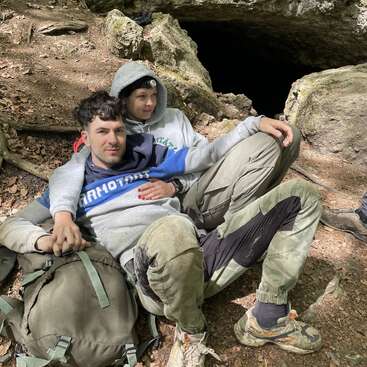 The image depicts a man and woman sitting together in front of a cave entrance, with the man wearing a backpack and the woman wearing a headlamp.
