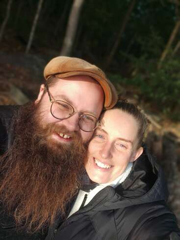 Um homem barbudo, usando óculos e boné, abraça uma mulher sorridente ao ar livre, sob a luz do sol. Eles parecem felizes juntos, cercados por árvores e pela natureza.