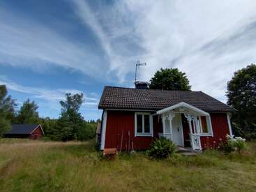 Un charmant cottage rouge avec des bordures blanches se trouve dans une cour herbeuse sous un ciel bleu avec des nuages ondulants, entouré d'arbres et d'un paysage de campagne paisible.