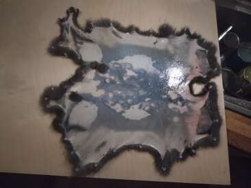 A wooden board with a large, irregular burnt patch in the center, surrounded by blackened edges, displaying charring and scorch marks from intense heat.