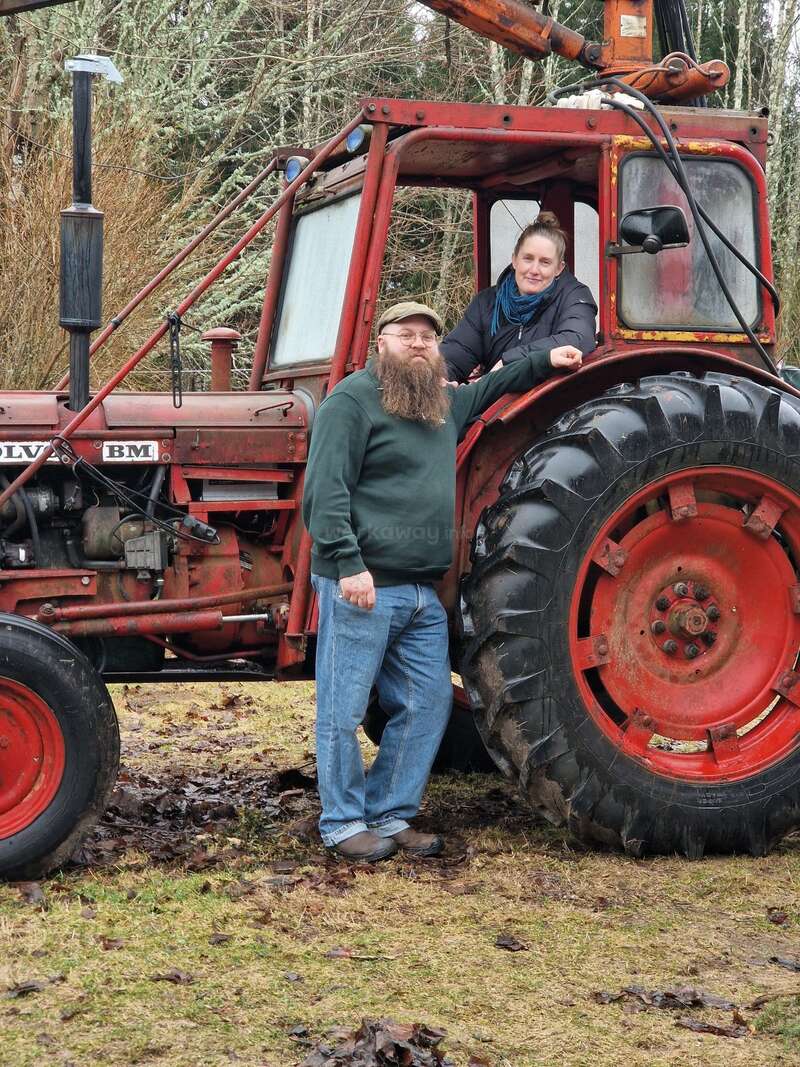 A bearded man stands by an old red tractor, while a woman sits inside. They both smile, surrounded by trees and muddy, grassy ground.