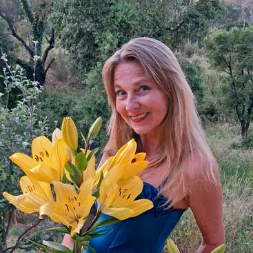 A smiling woman with long blonde hair stands outdoors, holding a bouquet of vibrant yellow lilies. Surrounded by lush greenery, she wears a blue dress, looking cheerful.