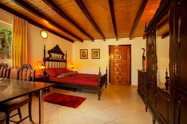 Dieses Zimmer hat eine Holzdecke, ein verziertes dunkles Bett, einen passenden Kleiderschrank, warme Beleuchtung, zwei Stühle, einen Tisch, einen gefliesten Boden, gelbe Vorhänge und gerahmte Kunstwerke an den Wänden.