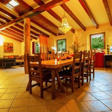 Uma sala de jantar aconchegante com uma mesa de madeira rústica, oito cadeiras, lustre, vigas expostas, piso de ladrilho, iluminação aconchegante e janelas grandes com vista para a paisagem verde ao ar livre.