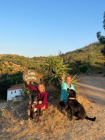 Duas mulheres e dois cães pretos sentados ao lado de uma placa "Casa Gaisma", posando alegremente em uma paisagem rural ensolarada, com colinas e vegetação ao fundo.