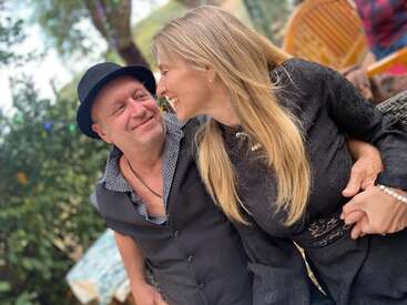 Um casal alegre senta-se junto ao ar livre, sorrindo e olhando nos olhos um do outro, segurando as mãos com carinho, cercado por vegetação e móveis de jardim casuais.