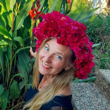 A woman with long blonde hair smiles brightly, wearing a vibrant crown of red flowers. She stands amid lush green plants and colorful garden surroundings, radiating happiness.
