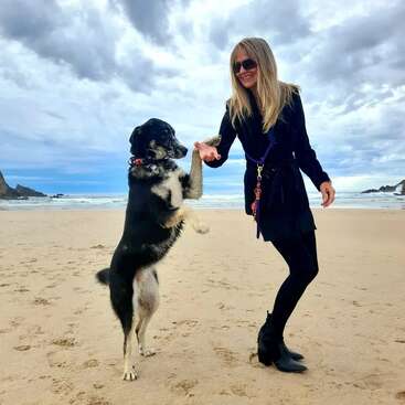 Uma mulher com roupas pretas sorri em uma praia de areia, de mãos dadas com um cachorro em pé. O céu está nublado e as ondas do mar são visíveis.