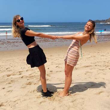 Duas mulheres se dão as mãos alegremente em uma praia ensolarada, sorrindo para a câmera. As ondas do mar e as pessoas distantes criam uma atmosfera de verão animada e despreocupada.
