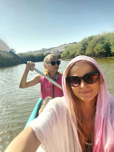 Duas mulheres andando de caiaque em um rio calmo, ambas usando óculos escuros. Uma rema enquanto a outra tira uma selfie, enrolada em um cachecol rosa claro, sob um céu claro.