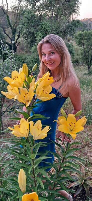 Uma mulher sorridente com longos cabelos loiros está ao ar livre, segurando um buquê de lírios amarelos vibrantes. Cercada por uma vegetação exuberante, ela usa um vestido azul, parecendo alegre.