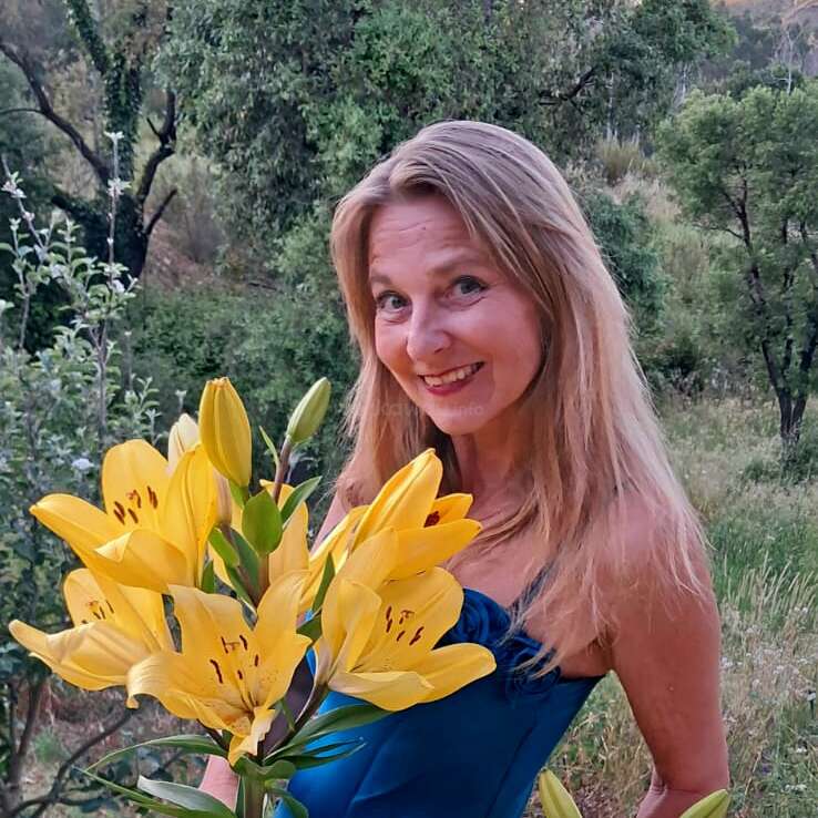 Eine lächelnde Frau mit langen blonden Haaren steht im Freien und hält einen Strauß leuchtend gelber Lilien in der Hand. Umgeben von üppigem Grün, trägt sie ein blaues Kleid und wirkt fröhlich.