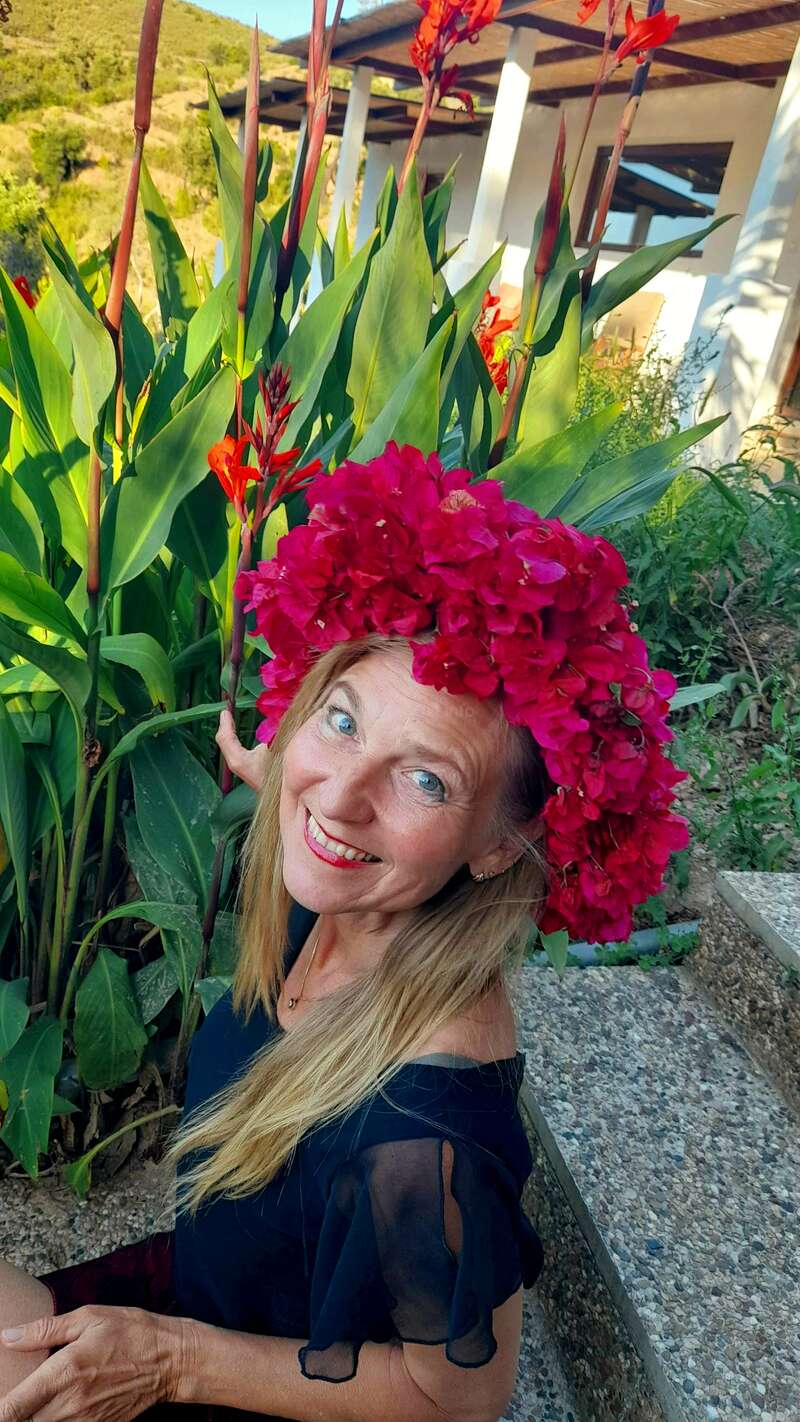 Uma mulher com longos cabelos loiros sorri brilhantemente, usando uma vibrante coroa de flores vermelhas. Ela está em meio a plantas verdes exuberantes e um jardim colorido, irradiando felicidade.