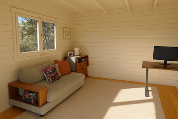 This cozy room features a light gray sofa with colorful pillows, a small wooden cabinet, a desk with a monitor, wooden floors, and natural sunlight.