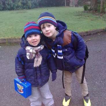 Das Bild zeigt zwei Jungen, die in Winterkleidung auf einer Straße stehen. Der eine hält eine blaue Lunchbox, der andere trägt einen Rucksack.