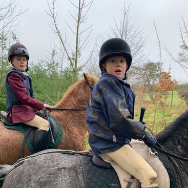 Das Bild zeigt zwei Kinder, die auf einem Feld reiten. Das eine Kind trägt eine blaue Jacke, das andere eine kastanienbraune Jacke und beide haben einen Helm auf.