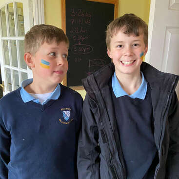 The image shows two boys standing side by side, wearing school uniforms, with a blackboard in the background featuring handwritten notes and a white door to the right.