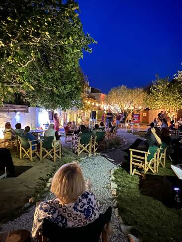 L'image représente une réunion animée en plein air dans une cour, avec une foule assise sur des chaises, entourée d'arbres et de bâtiments, sur fond de ciel bleu foncé.
