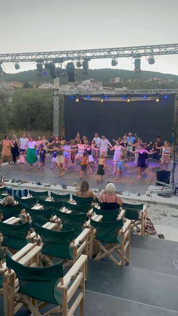 L'image représente un spectacle animé en plein air, avec une foule de personnes qui dansent sur scène et un public assis qui regarde depuis l'avant. La scène se déroule sur un fond pittoresque d'arbres et de maisons.