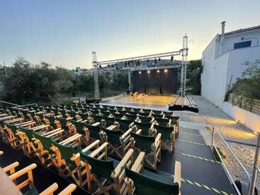 L'image représente un amphithéâtre en plein air avec une scène, des sièges et un chemin en gravier menant à la scène. Le gradin est équipé de chaises vertes de directeur.