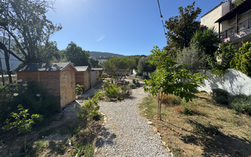 L'image représente une scène de jardin sereine, avec un chemin de gravier, des cabanes en bois et de la verdure luxuriante, sur fond de ciel bleu clair et de collines lointaines.