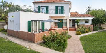 L'image représente une maison blanche à deux étages avec un toit en tuiles rouges, dotée d'un balcon, d'un patio et d'une verdure luxuriante dans la cour environnante.