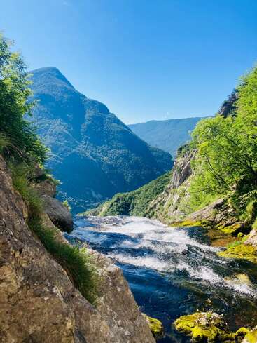 A vibrant mountain landscape features a cascading waterfall, lush green trees, clear blue sky, rugged rocks, and distant peaks, creating a breathtaking and serene natural scene.