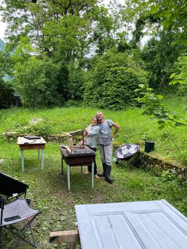 A smiling couple stands together in a lush green backyard with trees, surrounded by wooden tables and outdoor chairs, suggesting a relaxed, outdoor gathering or project.