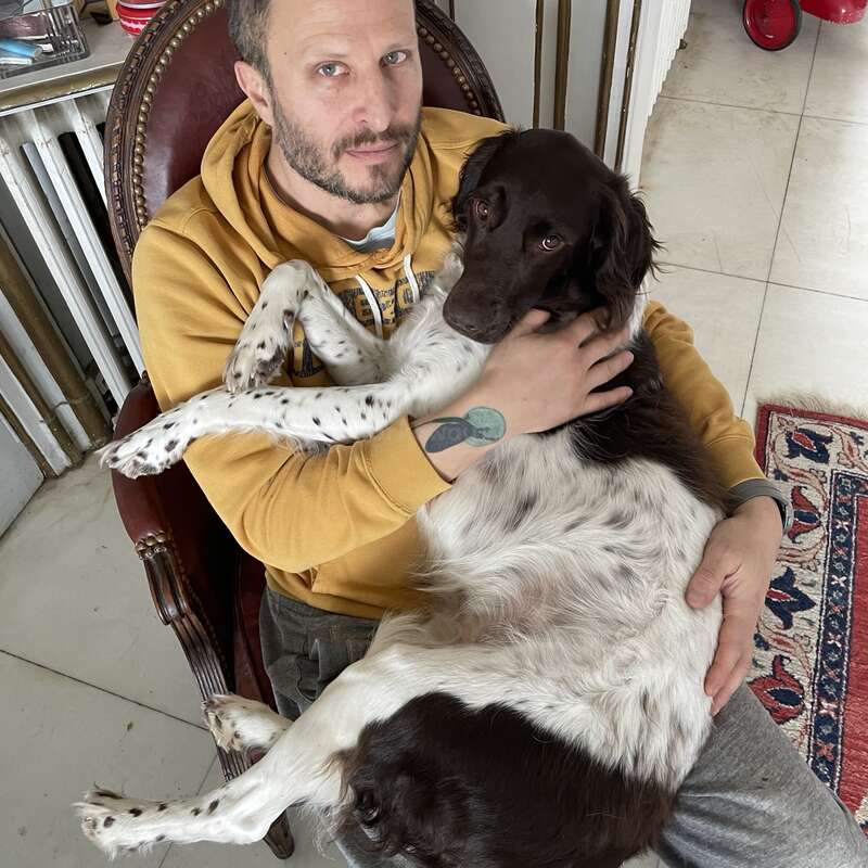 Ein Typ in einem senfgelben Kapuzenpulli sitzt auf einem Stuhl und hat einen großen braun-weißen Hund auf dem Schoß. Beide gucken ganz entspannt in die Kamera.