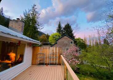 Une terrasse confortable donne sur un jardin avec des arbres en fleurs. Une cheminée brille à l'intérieur de la maison, tandis que le ciel coloré et la verdure luxuriante renforcent l'atmosphère sereine et accueillante.