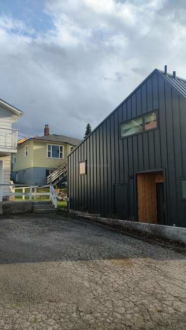 This image shows a modern black house with wooden accents beside older homes, cracked driveway pavement, white railings, greenery, and a cloudy sky above, suggesting a residential area.