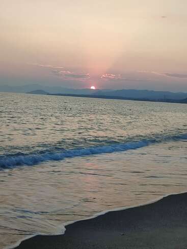 A serene beach scene at sunset; gentle waves kiss the shore, mountains outline the horizon, and the orange sun dips behind soft, pastel-colored clouds.
