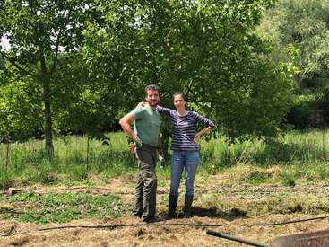 A man and woman stand together outdoors, surrounded by green trees and grass. The weather looks sunny. They appear happy, possibly working or gardening together.