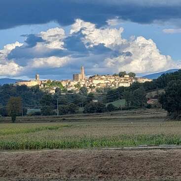 A picturesque hilltop village is illuminated by sunlight, nestled among trees and fields, under a dramatic sky filled with large, billowing clouds and contrasting shadows.