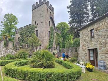 A stone castle tower with crenellations stands amid lush gardens and greenery. Two people tend to plants near a charming stone house adorned with vibrant flowers.
