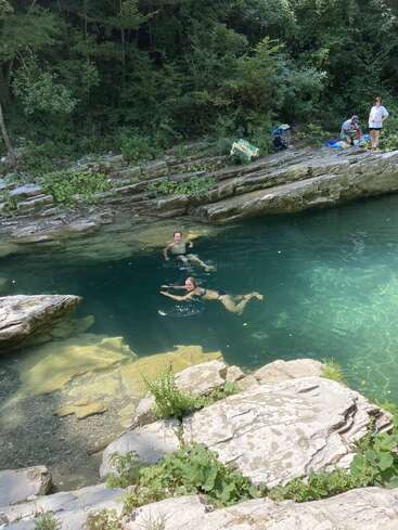 Two people happily swim in a clear, turquoise natural pool surrounded by rocks and lush greenery, while three others relax and observe from the rocky shore above.