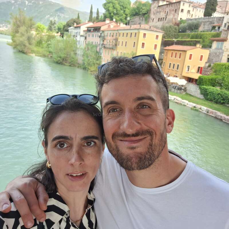 Um casal sorridente posa para uma selfie em um rio panorâmico, com prédios coloridos, vegetação exuberante e montanhas distantes criando um cenário europeu pitoresco e ensolarado.