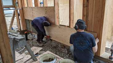Dois homens estão trabalhando em um canteiro de obras, construindo uma casa. Eles estão usando coletes de segurança e estão cercados por materiais de construção. A cena tem como pano de fundo um bairro suburbano.