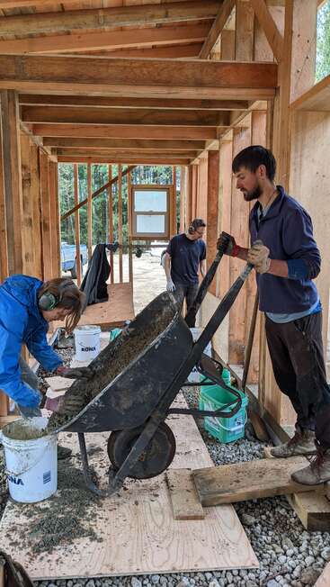 A imagem mostra três indivíduos envolvidos em trabalhos de construção em um edifício com estrutura de madeira, utilizando um carrinho de mão e baldes em meio a um cenário de árvores e um terreno coberto de cascalho.