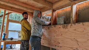 Dois homens estão construindo uma casa, um vestindo uma camisa amarela e o outro uma camisa branca. Eles estão em pé em uma plataforma de madeira, cercados por um corpo de água. A cena é serena e pacífica.