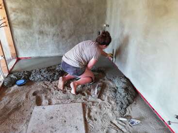 Uma mulher está ajoelhada no chão de uma sala em construção, trabalhando com cimento e ferramentas, vestindo camiseta e shorts.
