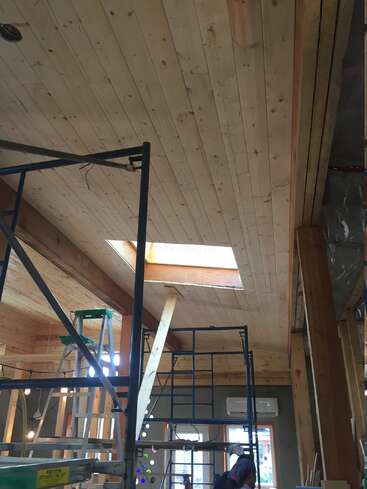 A imagem retrata uma sala em construção, com um teto de madeira com uma claraboia, andaimes e um trabalhador ao fundo, cercado por materiais de construção.
