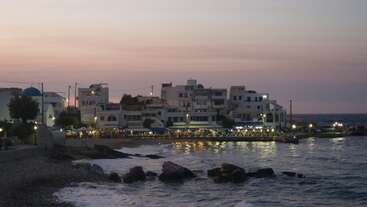 A serene coastal village at dusk, with white buildings, glowing streetlights, gentle waves, rocky shoreline, and a pink-hued sky creating a peaceful, inviting Mediterranean atmosphere.