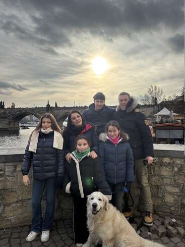 Una familia de seis miembros, entre los que se cuentan los niños y un perro, posa alegremente junto a un muro de piedra, con un pintoresco puente, un río y un cielo nublado de aspecto espectacular al fondo.