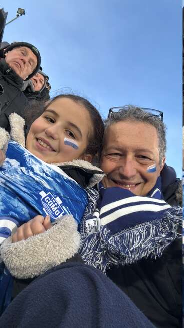 Un hombre sonriente y una niña, ambos con la cara pintada de azul y blanco y bien abrigados, asisten a un evento deportivo al aire libre para animar al equipo de fútbol del Como.