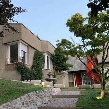Una casa moderna de arquitectura cúbica se alza entre árboles y arbustos frondosos, con grandes ventanales, una escalera de caracol roja, un camino de piedra y un ambiente de jardín tranquilo.