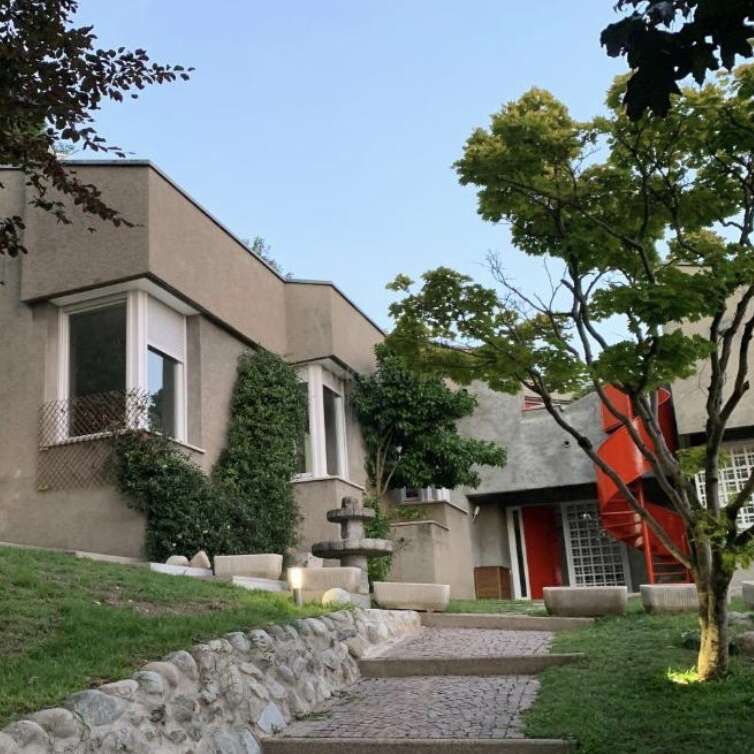 Una casa moderna de arquitectura cúbica se alza entre árboles y arbustos frondosos, con grandes ventanales, una escalera de caracol roja, un camino de piedra y un ambiente de jardín tranquilo.